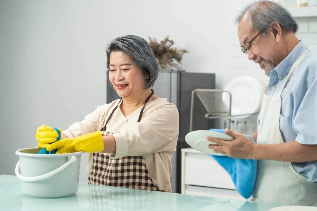 Two older adults wash and dry dishes together in a kitchen; one wears yellow gloves and uses a towel, while the other dries a plate with a blue cloth.