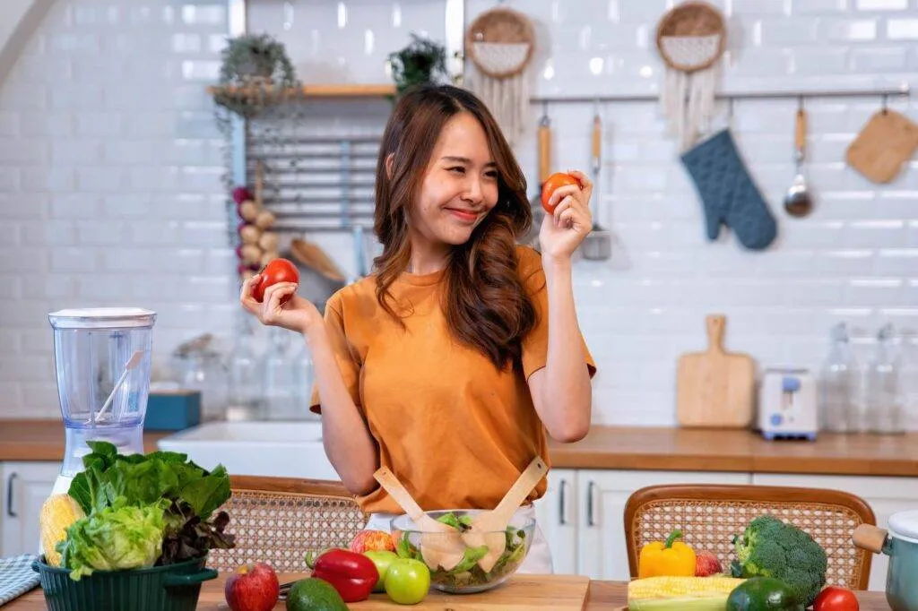 A woman in an orange shirt stands in a kitchen holding two tomatoes, surrounded by various fresh vegetables and a salad bowl on the table.