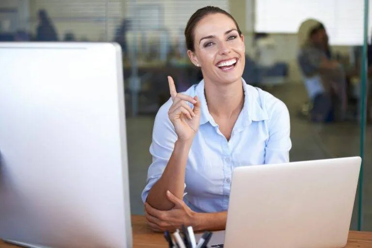 Woman in a blue shirt smiles and raises her index finger while sitting at a desk with two computers in an office setting.
