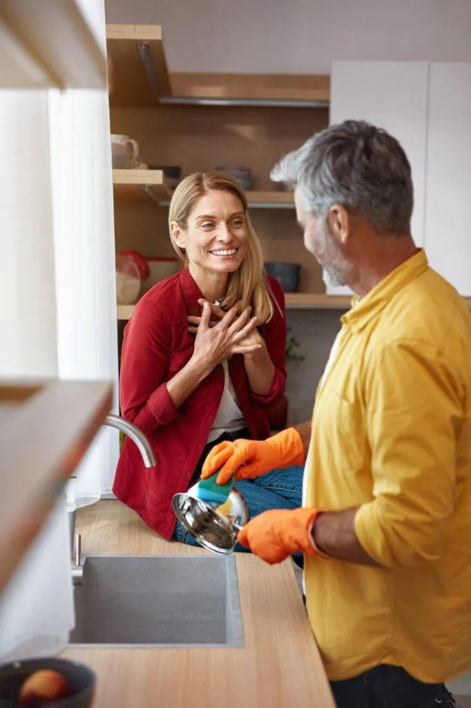 A man in orange gloves washes a pot at the kitchen sink, while a smiling woman in a red shirt stands nearby with her hands on her chest.
