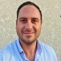 Man with short dark hair and light facial hair wearing a white shirt with blue dots stands in front of a textured beige wall in Phoenix, smiling at the camera, ready to enjoy office coffee and healthy snacks.
